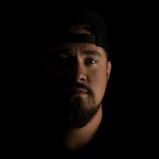 A man with a beard and mustache wears a backward black cap, partially lit with shadow covering most of his face against a black background.