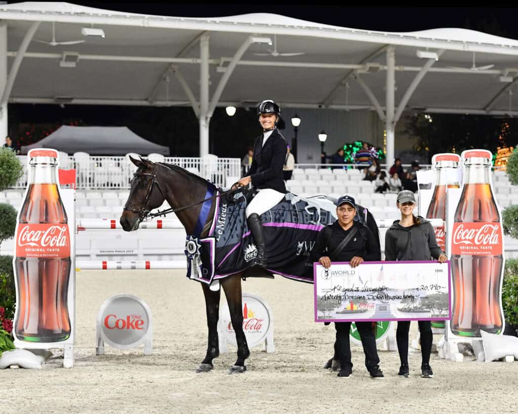 A rider on a horse poses with two people holding an oversized check in an arena, flanked by large Coke bottle displays and bleachers in the background.