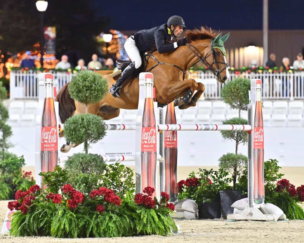 A rider on a brown horse in mid-jump over an obstacle decorated with Coca-Cola bottles at an equestrian event.