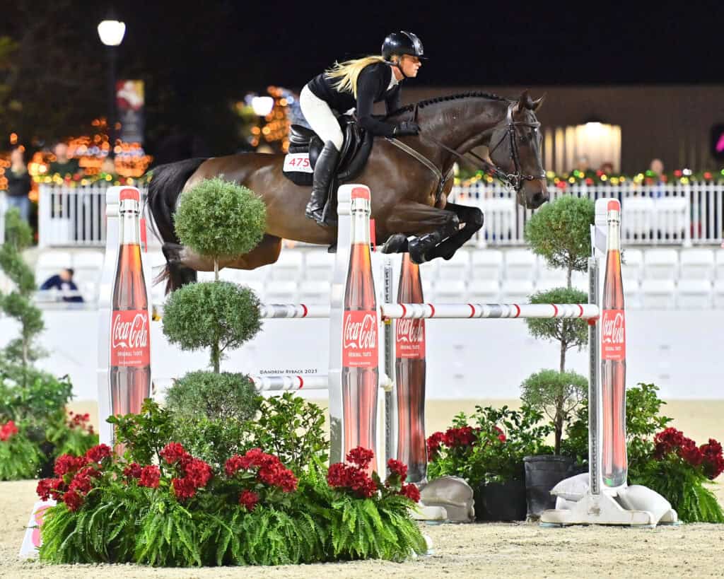 A rider in equestrian gear guides a horse over a jump decorated with Coca-Cola branding, flowers, and greenery at an outdoor night event.