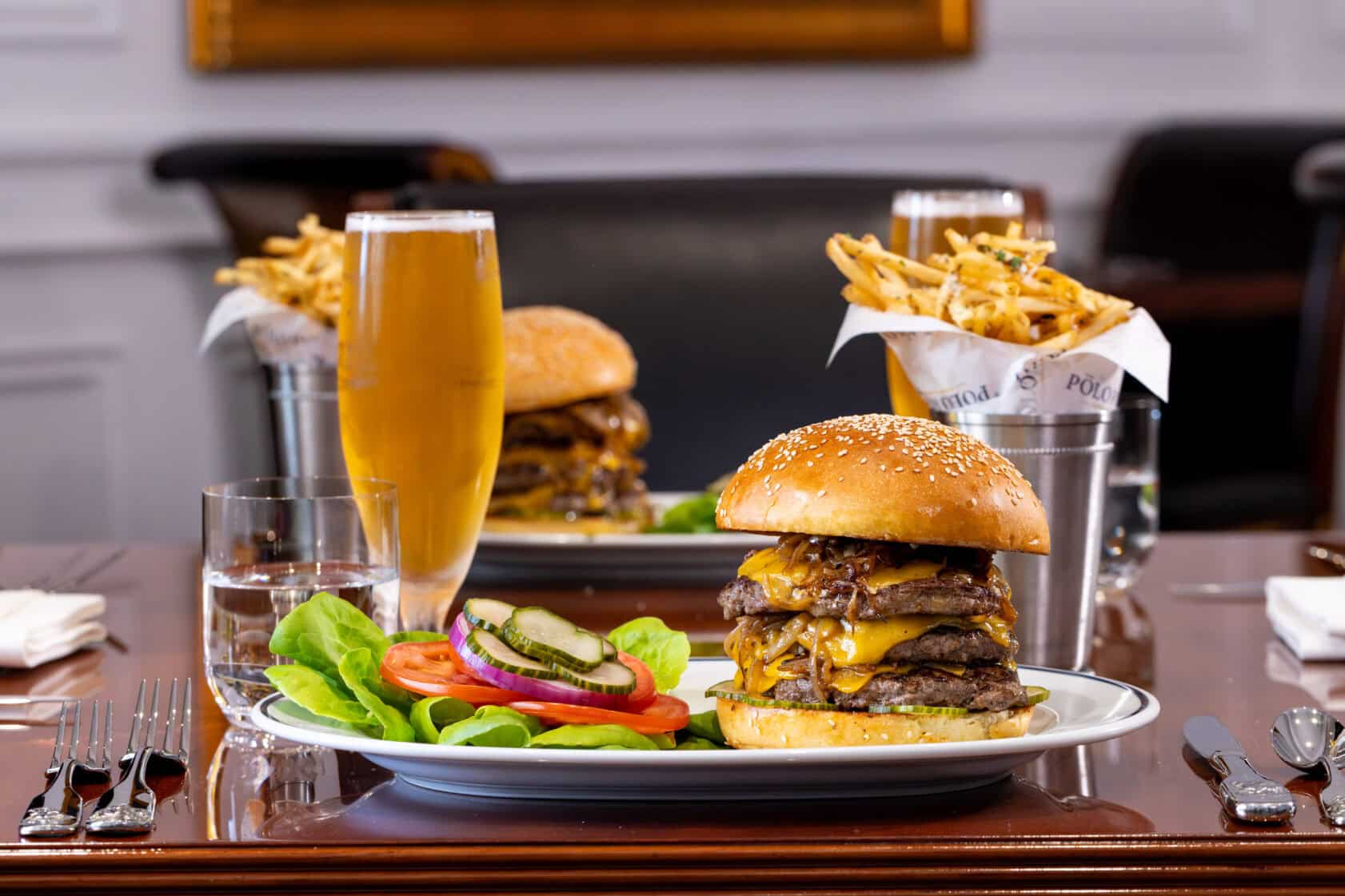 A large cheeseburger with lettuce, tomato, onion, and pickles is served on a plate with fries and a glass of beer on a restaurant table.