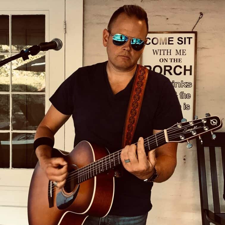 A man wearing sunglasses and a black t-shirt plays an acoustic guitar in front of a microphone, with a sign in the background inviting people to sit on the porch.