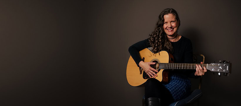 A woman with long curly hair sits on a chair, smiling and playing an acoustic guitar against a plain dark background.