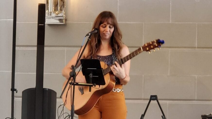 A woman plays an acoustic guitar and sings into a microphone, standing in front of a beige tiled wall.