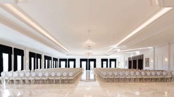 A large, elegant ballroom with marble floors, rows of white chairs with gold trim, chandeliers, and tall windows with black curtains.