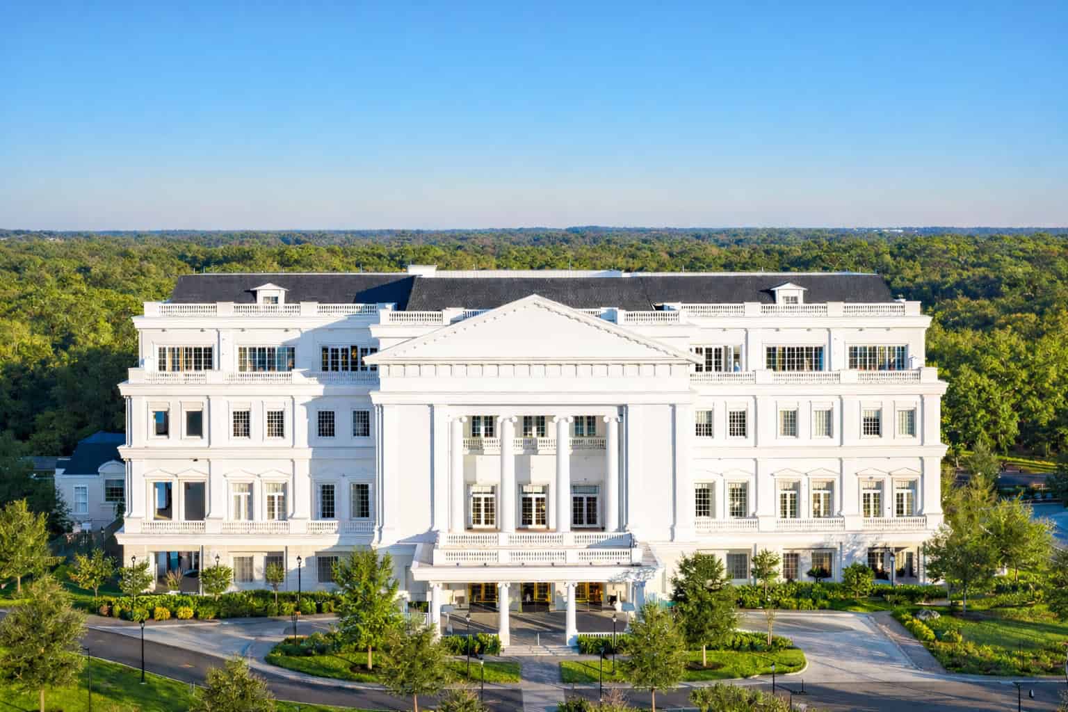 A large white neoclassical building with columns and multiple windows, surrounded by trees under a clear blue sky.
