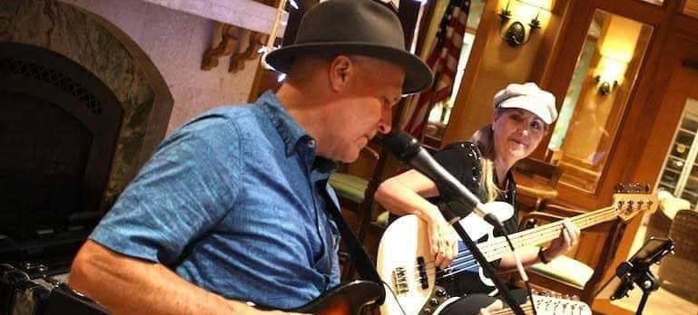 Two musicians play guitars indoors; one is singing into a microphone while the other looks on and plays a bass guitar.