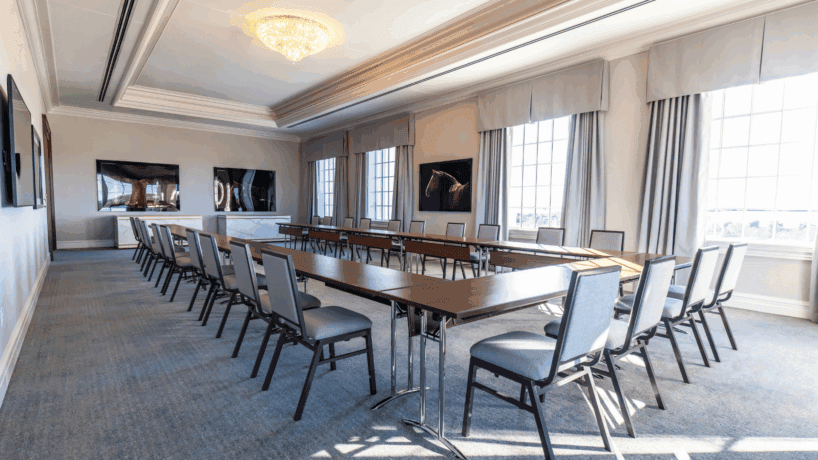 A modern conference room with U-shaped tables, gray chairs, large windows letting in natural light, and framed artwork on the walls.