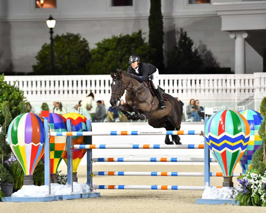 A person riding a horse jumps over a colorful obstacle in an equestrian competition, with hot air balloon decorations on both sides.