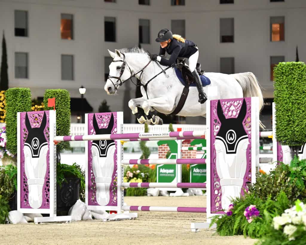 A rider on a white horse jumps over a pink and white obstacle during an equestrian event, with a building visible in the background.