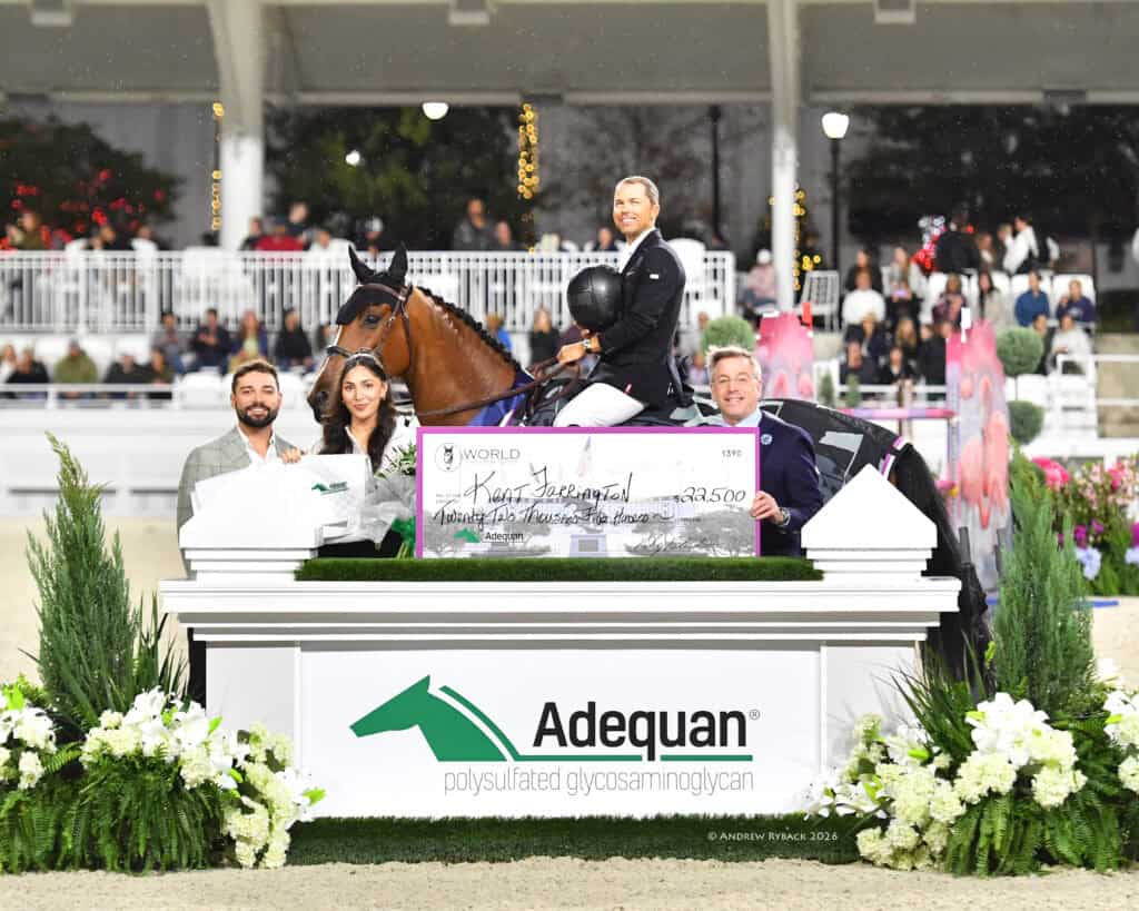 A rider on horseback holds an oversized check, flanked by three people, in front of an Adequan-branded display at an equestrian event.