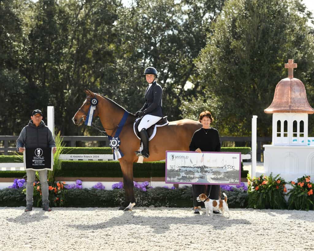 A rider on a horse poses with two people, one holding a trophy and the other holding a large check, next to a dog, at an outdoor equestrian event.