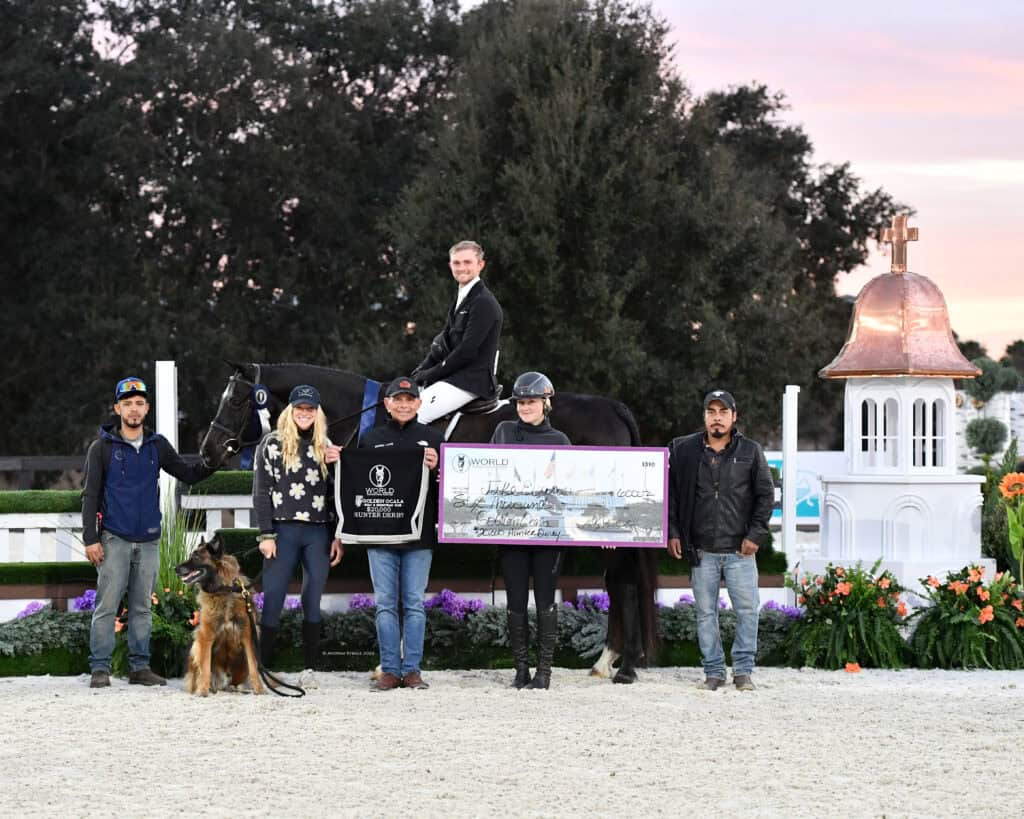 A group of six people and a dog pose outdoors; one person is on a black horse, another holds a large check, with trees and flowers in the background.