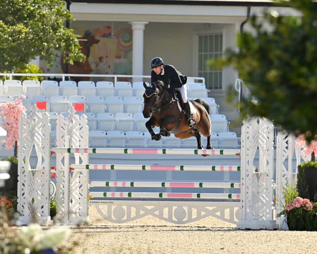 Equestrian rider and horse jump over a hurdle during a show jumping competition in an outdoor arena with empty white seats in the background.