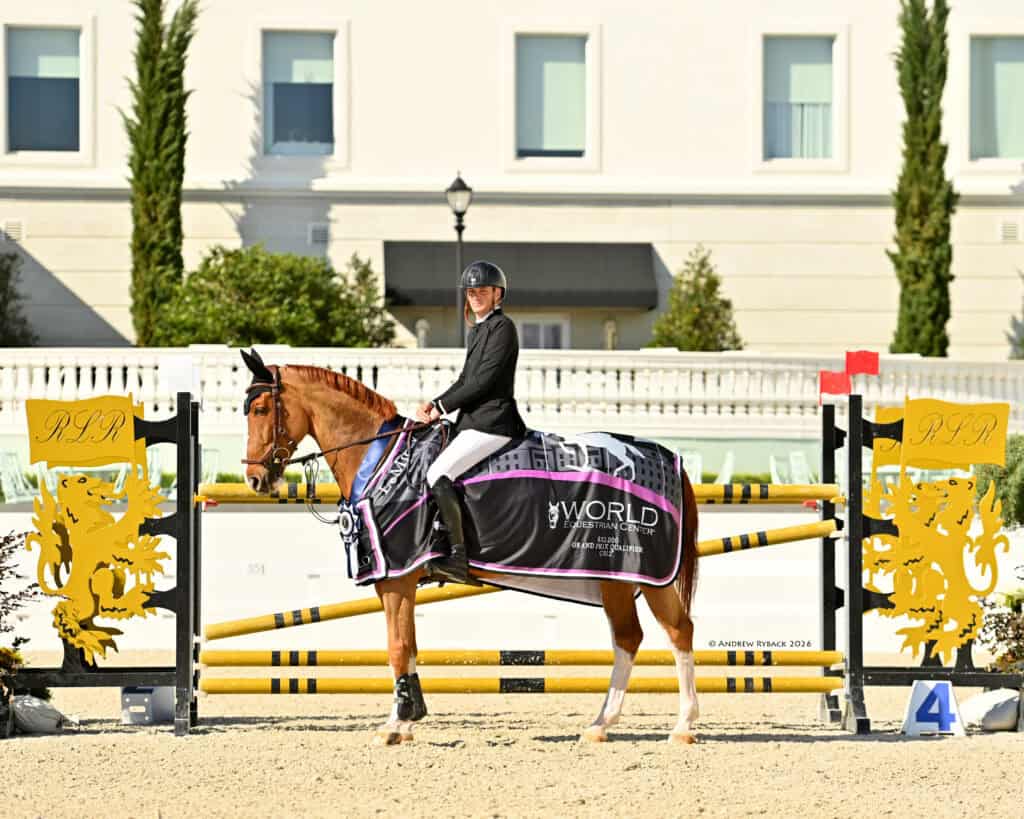 A rider in equestrian attire sits on a horse draped with a winner's blanket in front of show jumping obstacles at an outdoor arena.