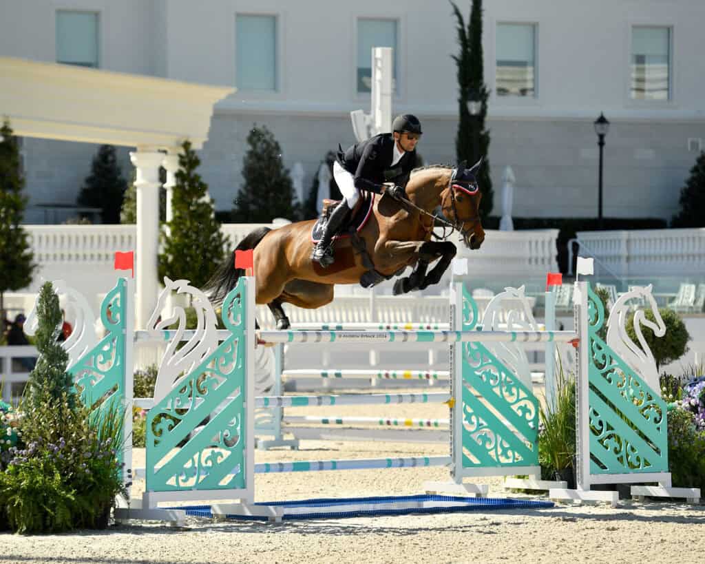 An equestrian and horse jump over a decorative obstacle during a show jumping competition in an outdoor arena.