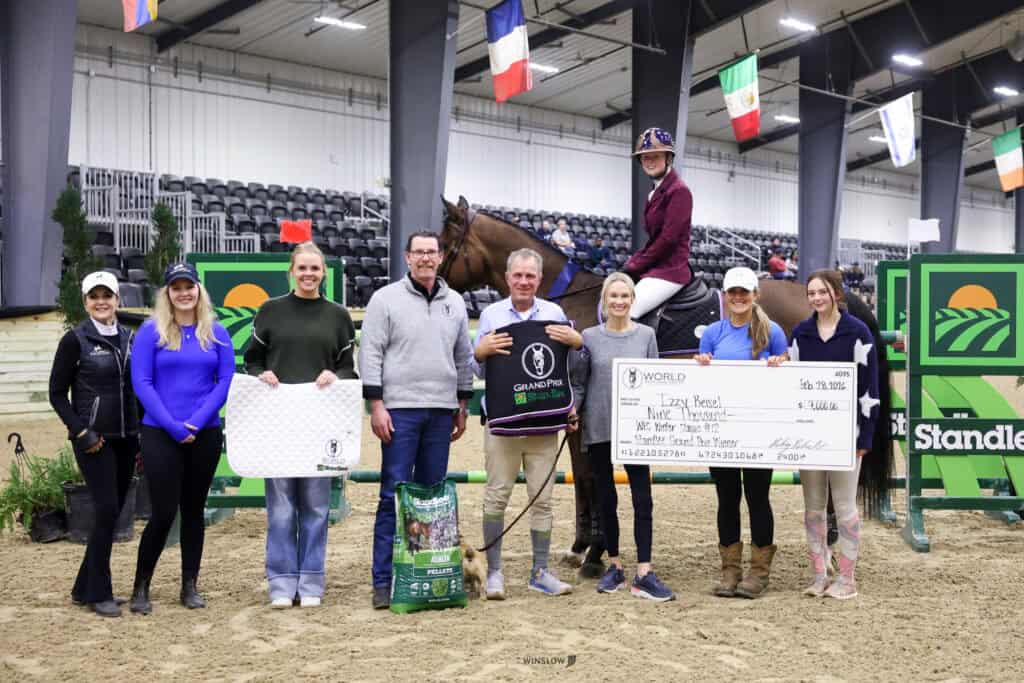 A group of people stands indoors on a sandy arena with a horse and large prize check; equestrian jumps and flags are visible in the background.