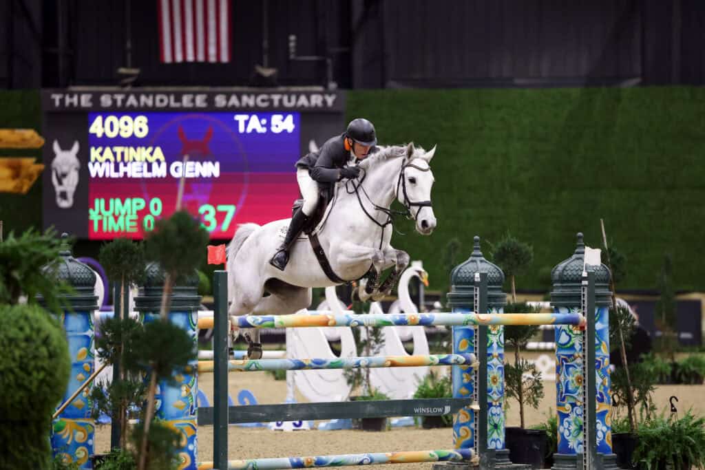 A rider on a white horse jumps over a colorful obstacle during an indoor equestrian event, with a scoreboard and greenery in the background.