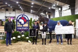 A group of people and a horse pose indoors with a large check, a trophy, and a Hagyard Equine Medical Institute sign at an equestrian event.