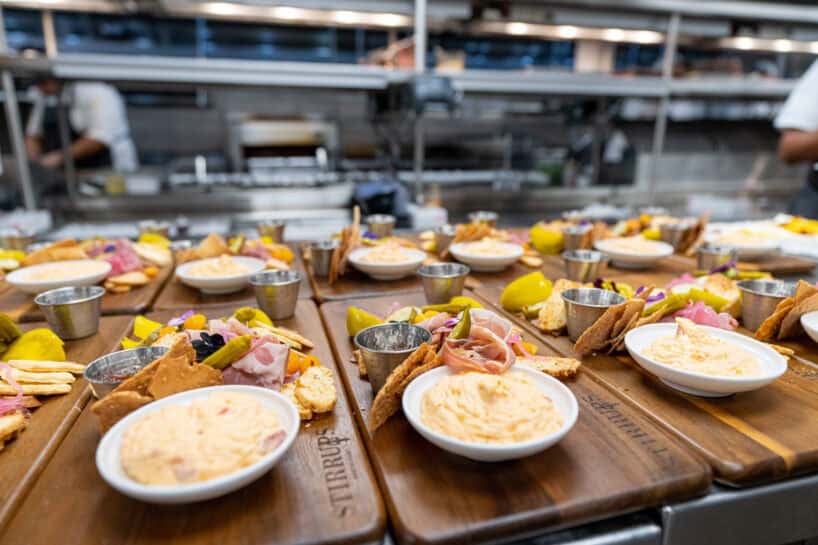 Several wooden boards topped with small dishes of dip, crackers, pickles, pita bread, and sliced meat are arranged on a kitchen counter, with a busy kitchen visible in the background.