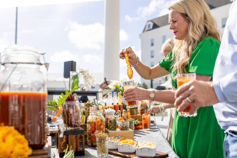 A woman in a green dress prepares a drink at an outdoor bar with various garnishes and ingredients on the counter in daylight.