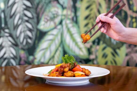 A hand holding chopsticks picks up a piece of stir-fried chicken and vegetables from a white plate, with a leafy wallpaper background.