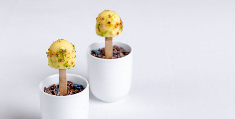 Two white cups filled with cocoa nibs hold wooden sticks topped with yellow-coated confections, garnished with green and red sprinkles, on a white surface.