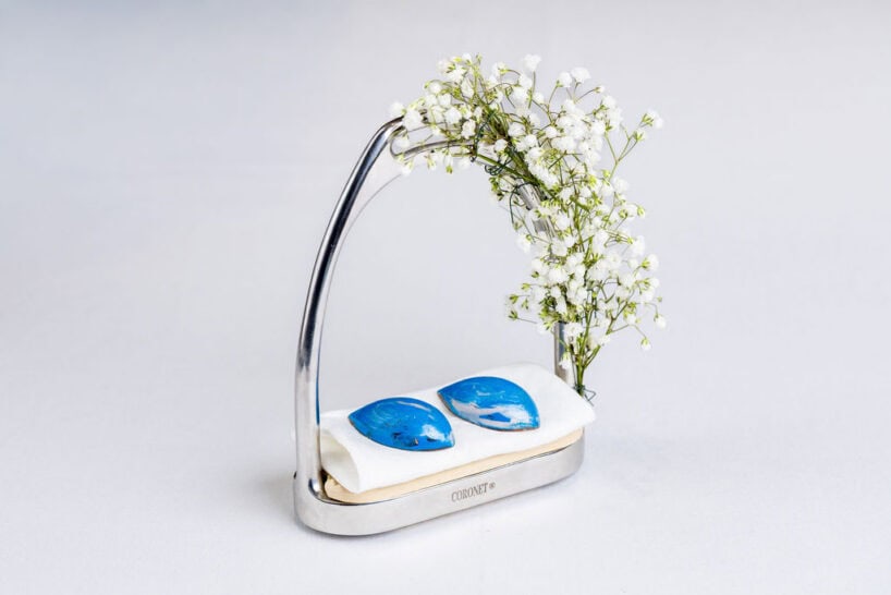 A silver stand holds two blue stones on a white cushion, adorned with small white flowers on the upper right side.