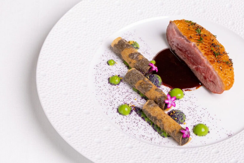 A plated dish featuring a seared piece of duck breast with sauce, decorative green and purple purees, edible flowers, and two rolled crisps on a white textured plate.