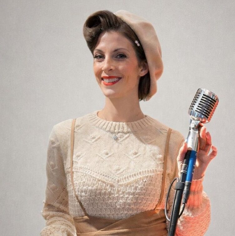 Woman in vintage-style clothing and beret stands smiling while holding a retro microphone against a plain background.
