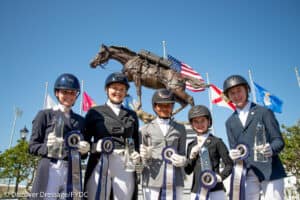 Five equestrian athletes in riding attire pose with ribbons and trophies in front of a horse statue and several national flags.