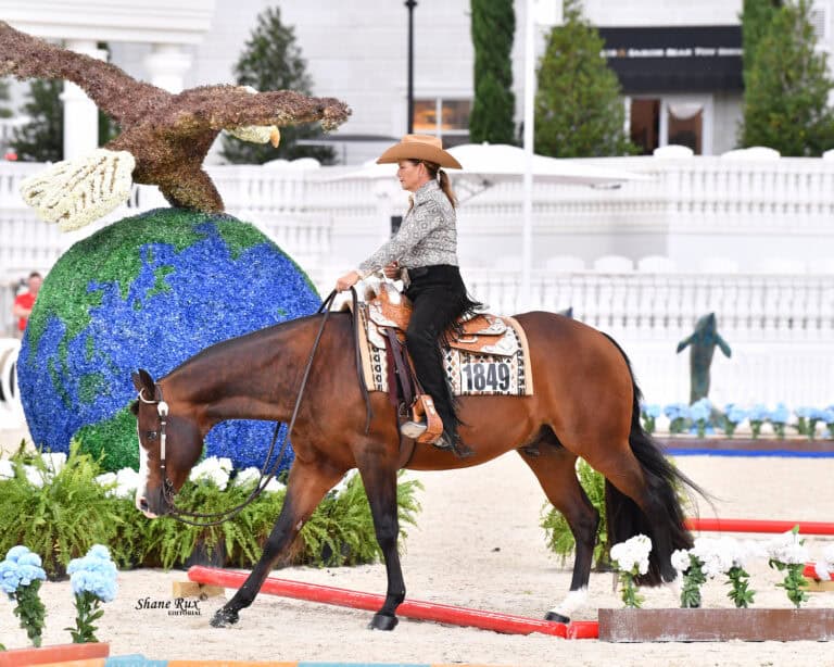 A woman in western attire rides a brown horse in an arena with decorative blue globe and eagle sculptures in the background.