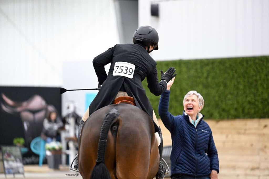 Equestrian rider on horseback, wearing number 7539, gives a high-five to a smiling woman standing beside the arena.