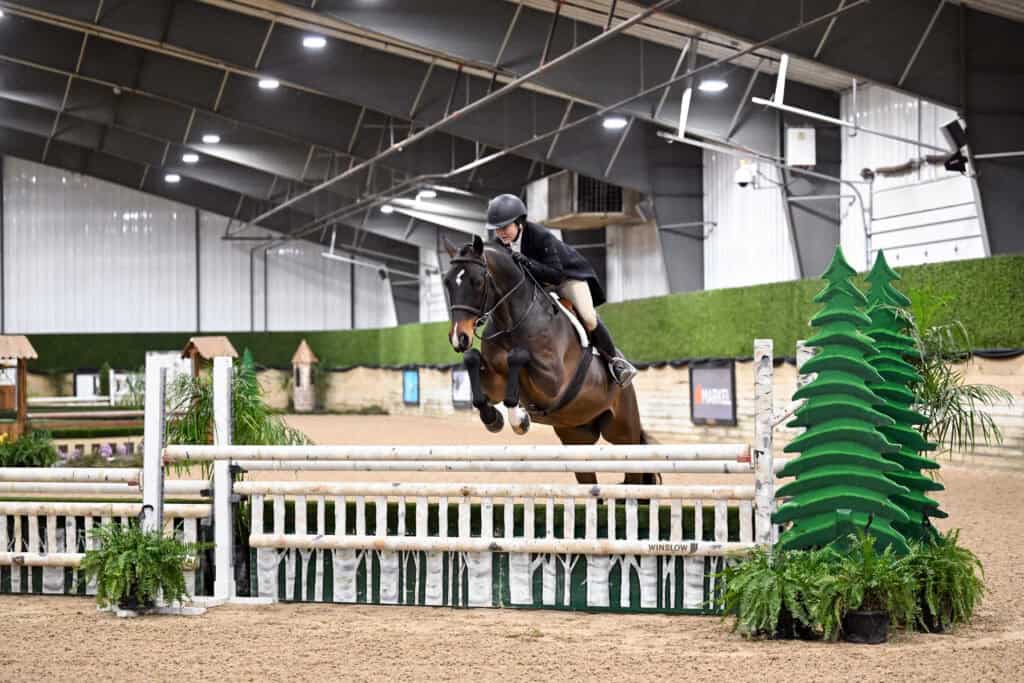A rider wearing a helmet and jacket jumps a brown horse over a white fence inside an indoor equestrian arena.
