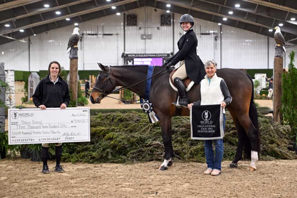 Three people pose in an indoor equestrian arena; one sits on a horse, another holds a large check, and the third holds a ribbon and plaque.