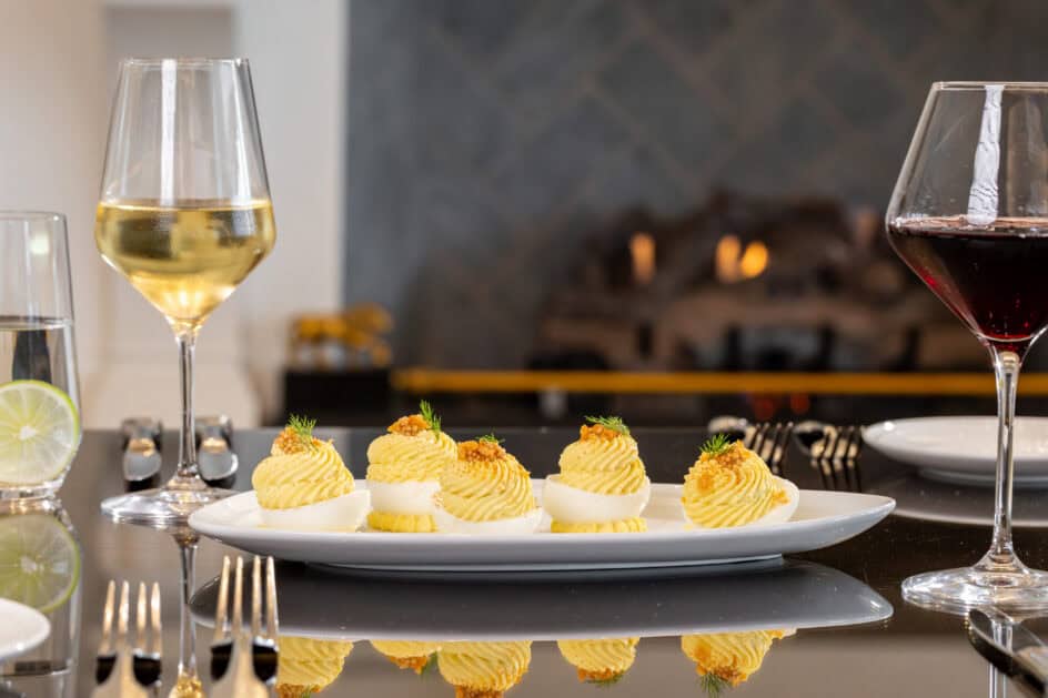 A plate of deviled eggs garnished with herbs is set on a table with wine glasses, silverware, and a fireplace in the background.