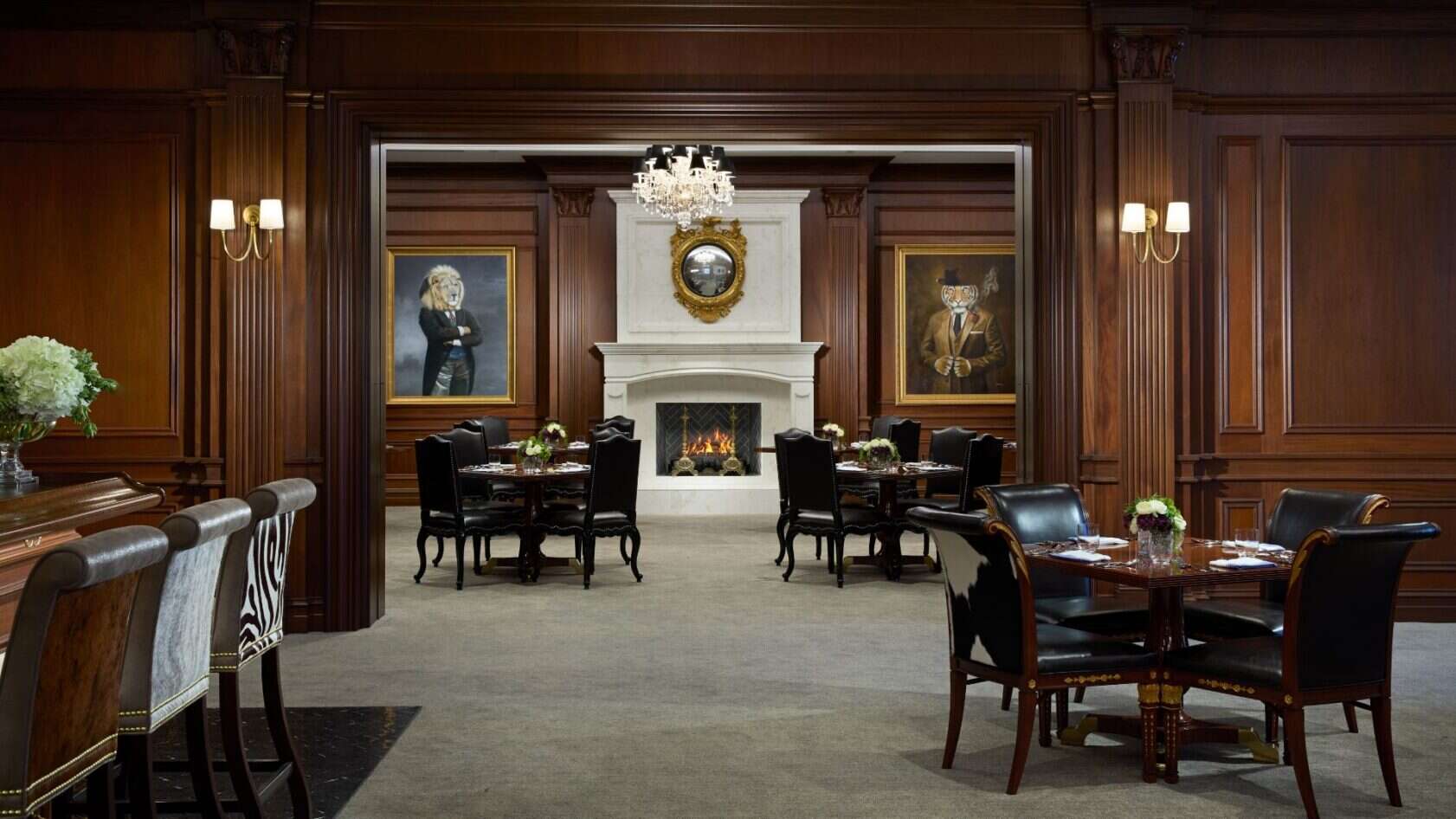 Elegant dining area with dark wood paneling, black leather chairs, tables set for service, a lit fireplace, chandelier, and two large portraits on either side of a mantel mirror.