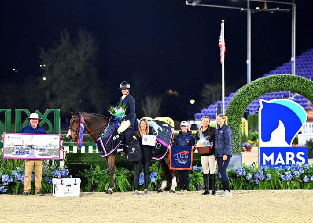 A group of people and a horse pose for a photo at an equestrian event award ceremony, with trophies, ribbons, a large check, and an event sponsor sign visible.