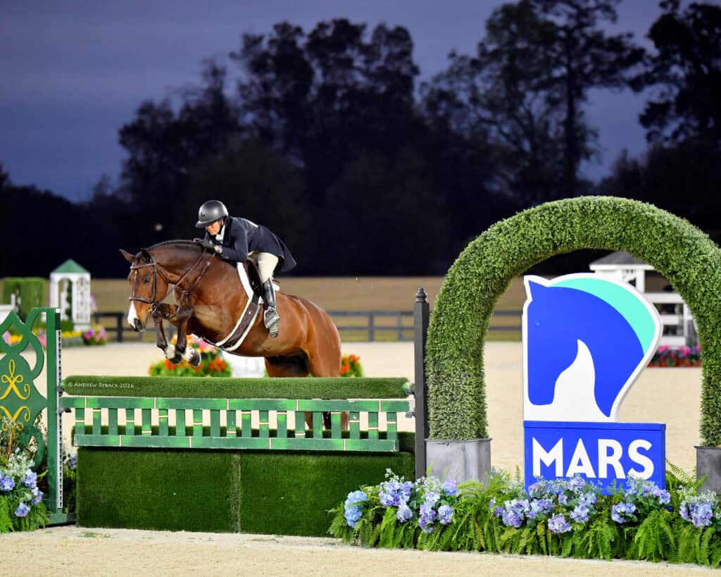 Equestrian rider and horse jumping over a green hedge fence at a competition, with a MARS logo sign and decorative flowers in the foreground.