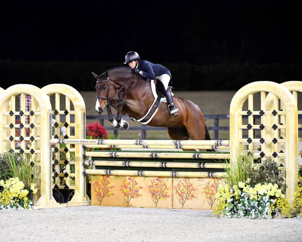 Equestrian rider in a helmet and gear jumps a horse over a yellow and green obstacle during a nighttime competition.