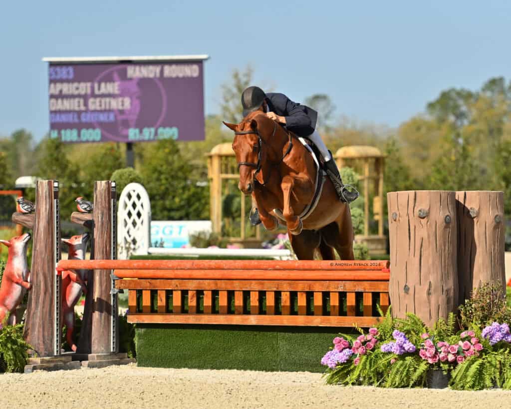 A rider on a horse jumps over an orange fence during an equestrian competition, with a scoreboard visible in the background.