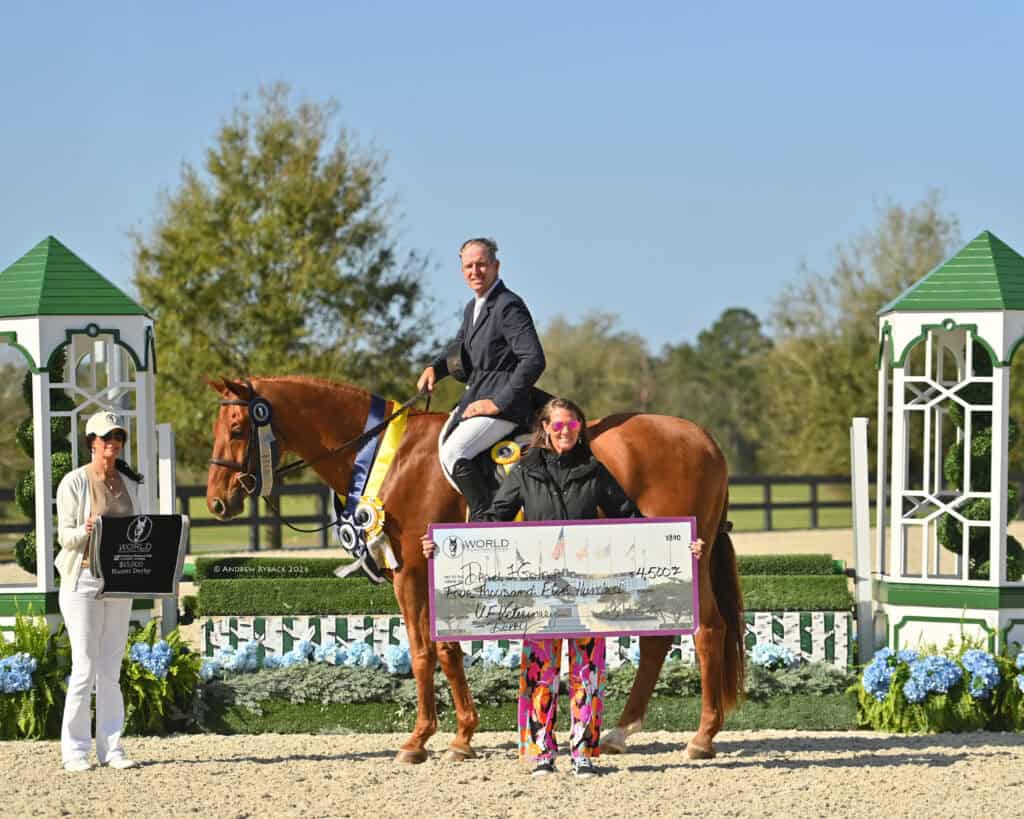 A man on a horse poses with two women, one holding a large check and another holding a plaque, in an outdoor equestrian awards setting.