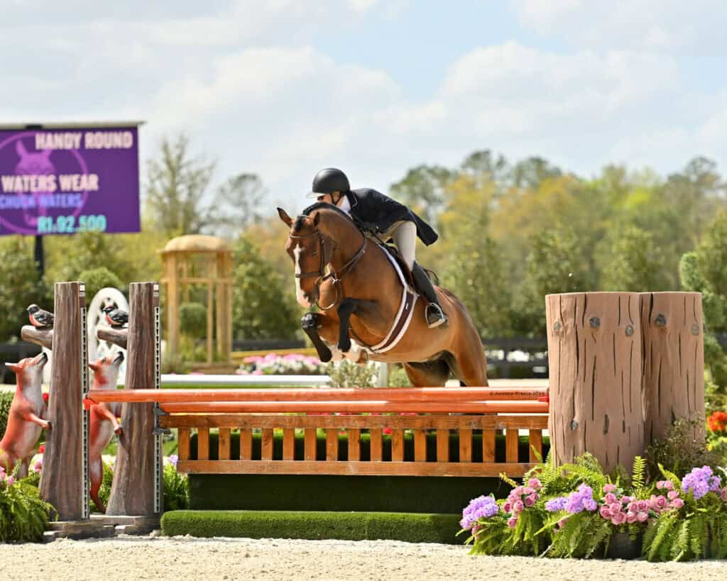 A rider in formal equestrian attire guides a horse over a jump in an outdoor arena decorated with flowers and wooden elements.