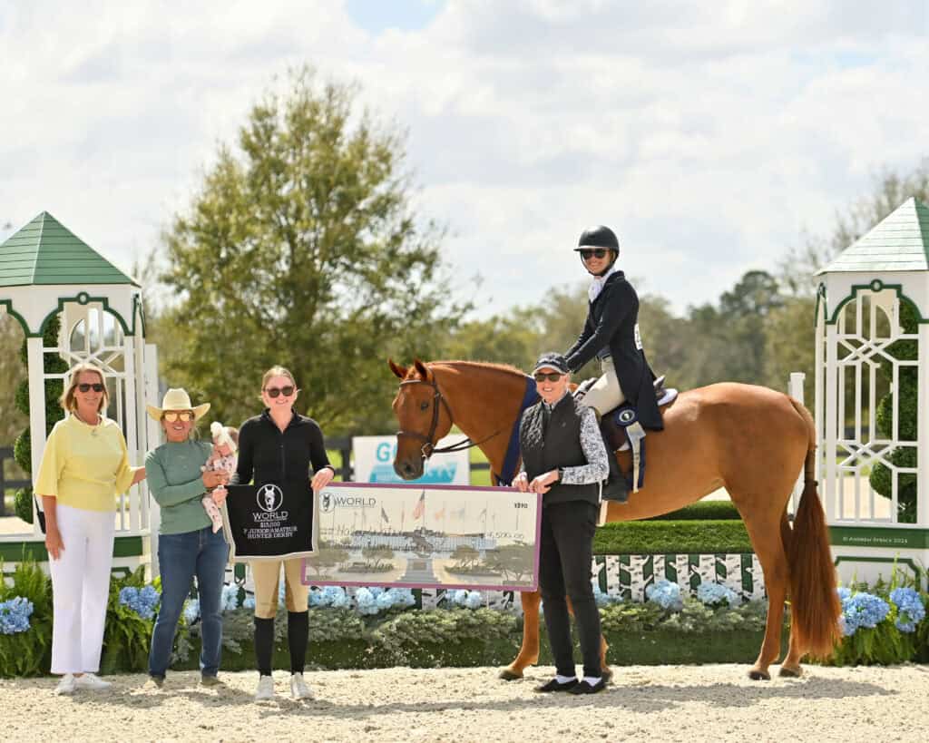 A group of five people and a horse pose outdoors; one person on horseback holds a trophy while another holds a large prize check.