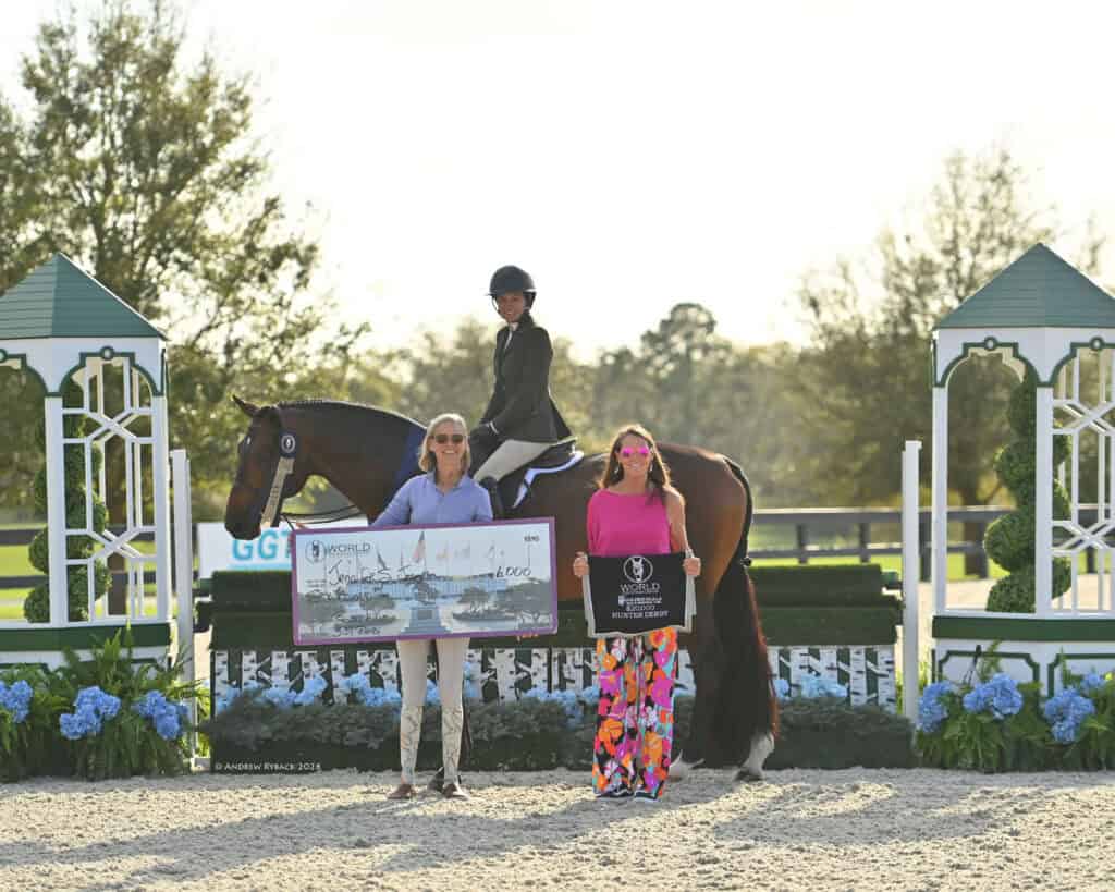 Three people at an outdoor equestrian event: one person on horseback, two women standing in front holding a large check and a plaque, with decorated white and green structures in the background.