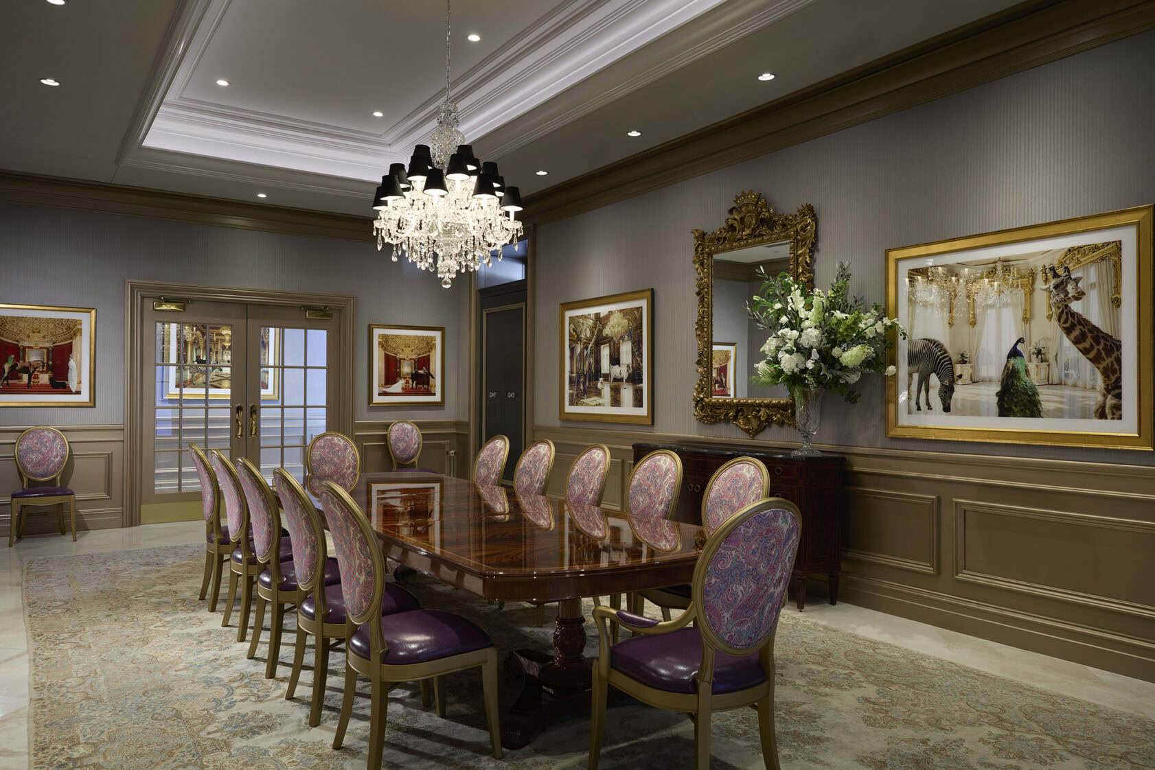 Elegant conference room with a polished wooden table, ornate chandelier, purple-upholstered chairs, large mirror, framed artwork, and floral arrangement on a patterned rug.