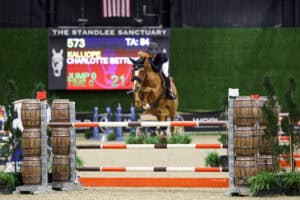 A rider and horse jump over an obstacle during an indoor equestrian event, with a scoreboard and barrels visible in the background.