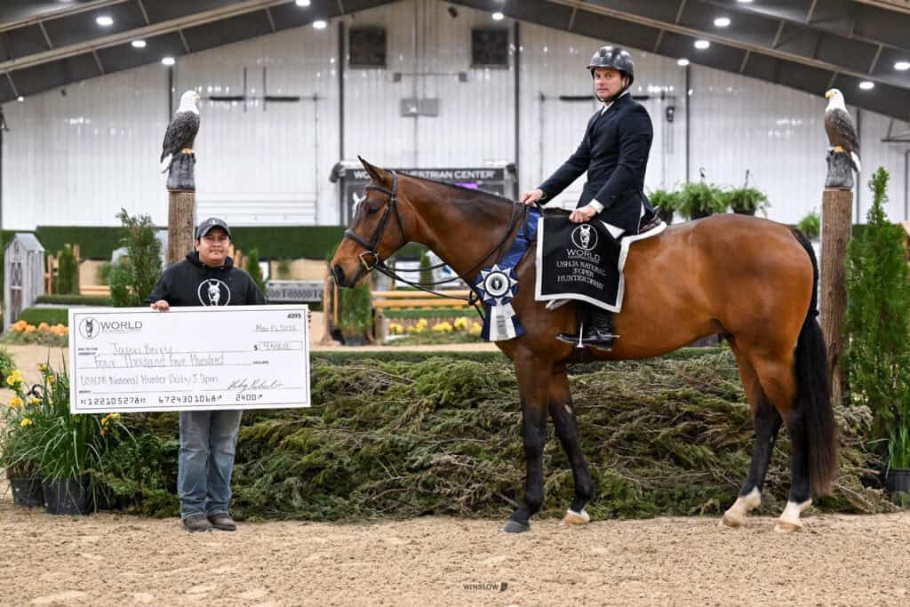 A man on horseback with a winner’s sash poses next to a man holding a large ceremonial check for $3,000 at an indoor equestrian event.