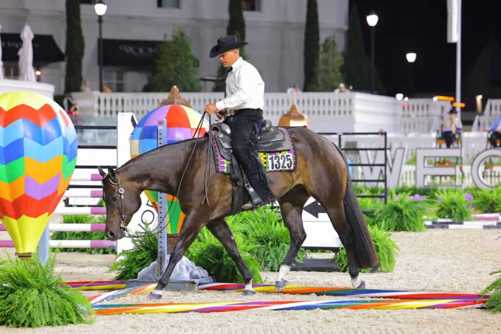 A rider in a white shirt and black hat rides a brown horse through a colorful obstacle course with balloon decorations at an equestrian event.