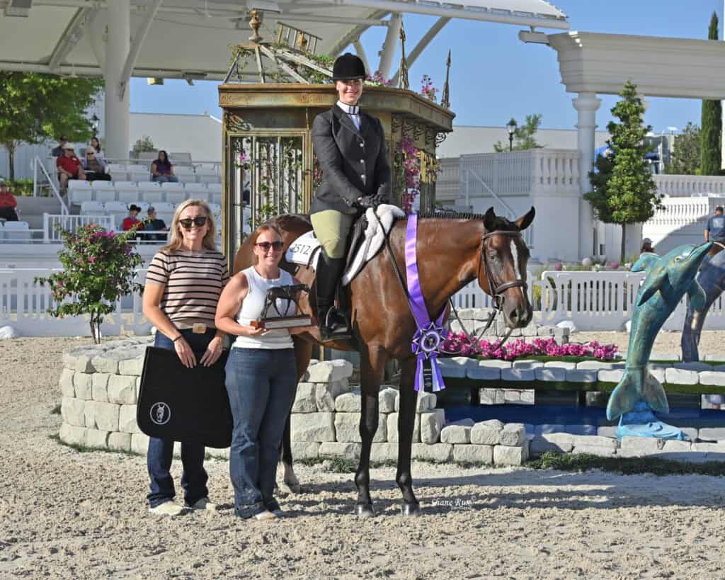 Three people pose with a horse wearing a purple ribbon; one woman is mounted, two stand beside, with a decorative gazebo and dolphin statues in the background.